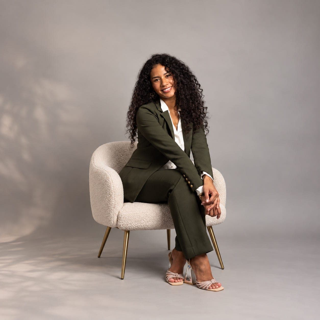 Nicol Rios seated in a cream chair, wearing a green suit, in a studio portrait.